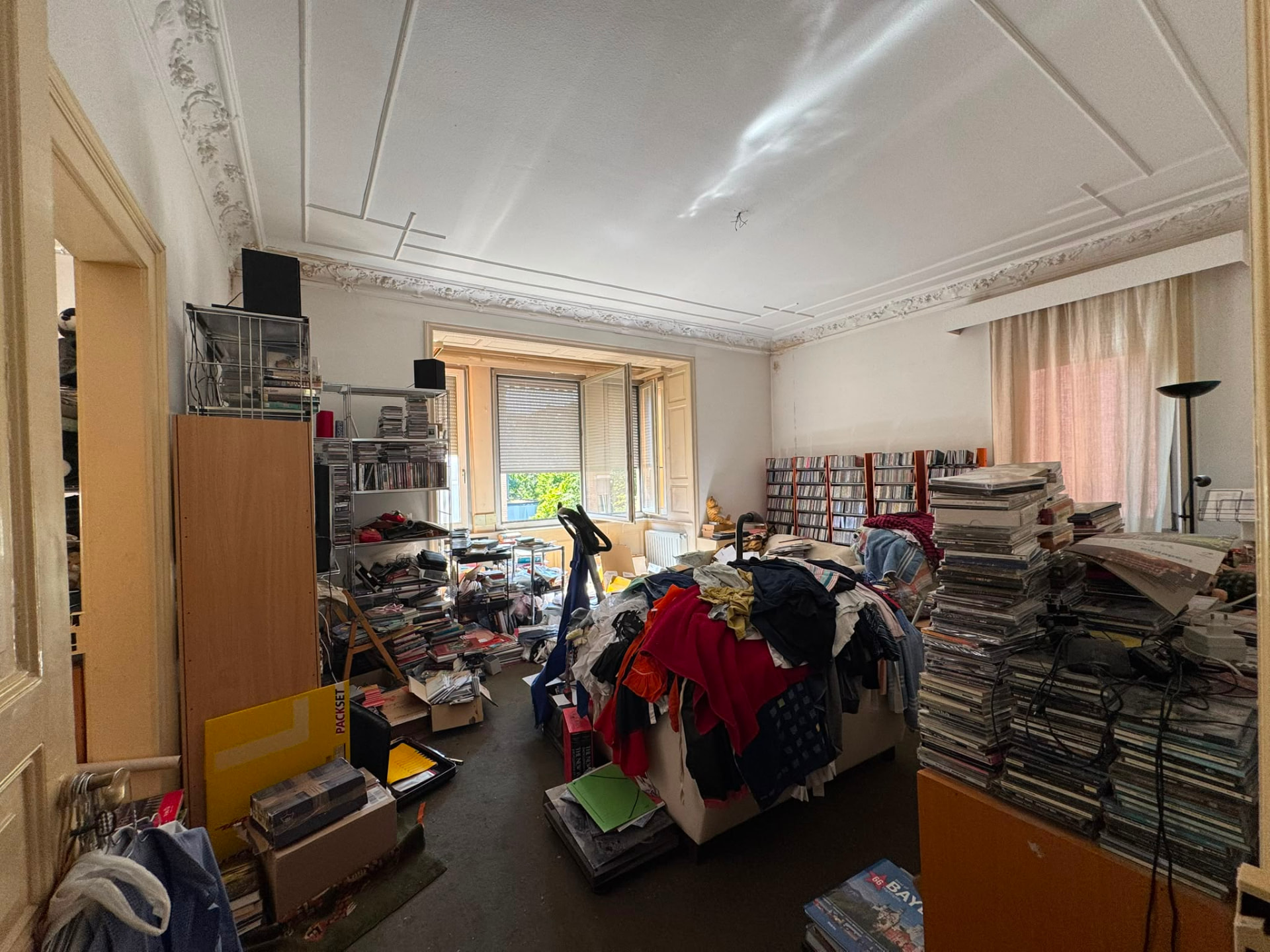 Vorher: mit CDs, Zeitschriften und Kleidung vollgestelltes Wohnzimmer einer Altbauwohnung vor der Entrümpelung